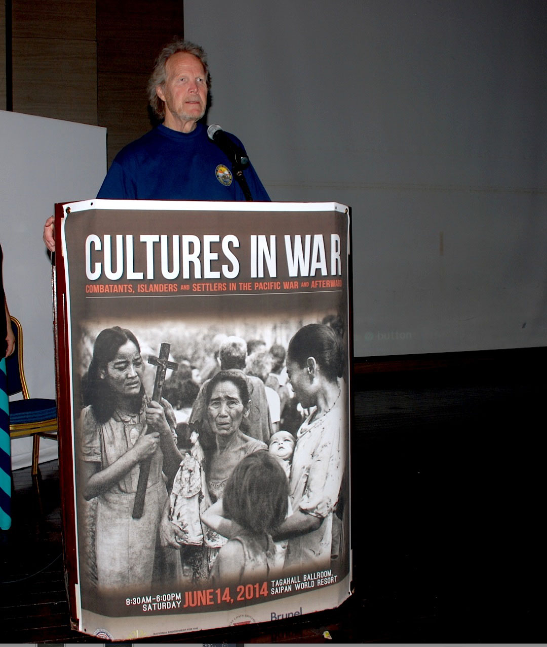 Photo of Anderson Giles speaking at a podium with poster for "Cultures in War" on the front